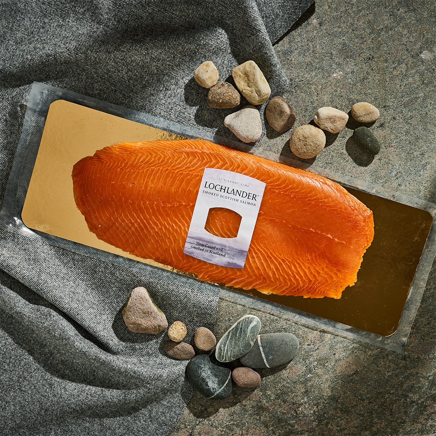 A large orange salmon fillet on a gold rectangular tray, with a white Lochlander smoked salmon label attached. The tray is styled on a gray stone surface with draped gray fabric and scattered river stones.