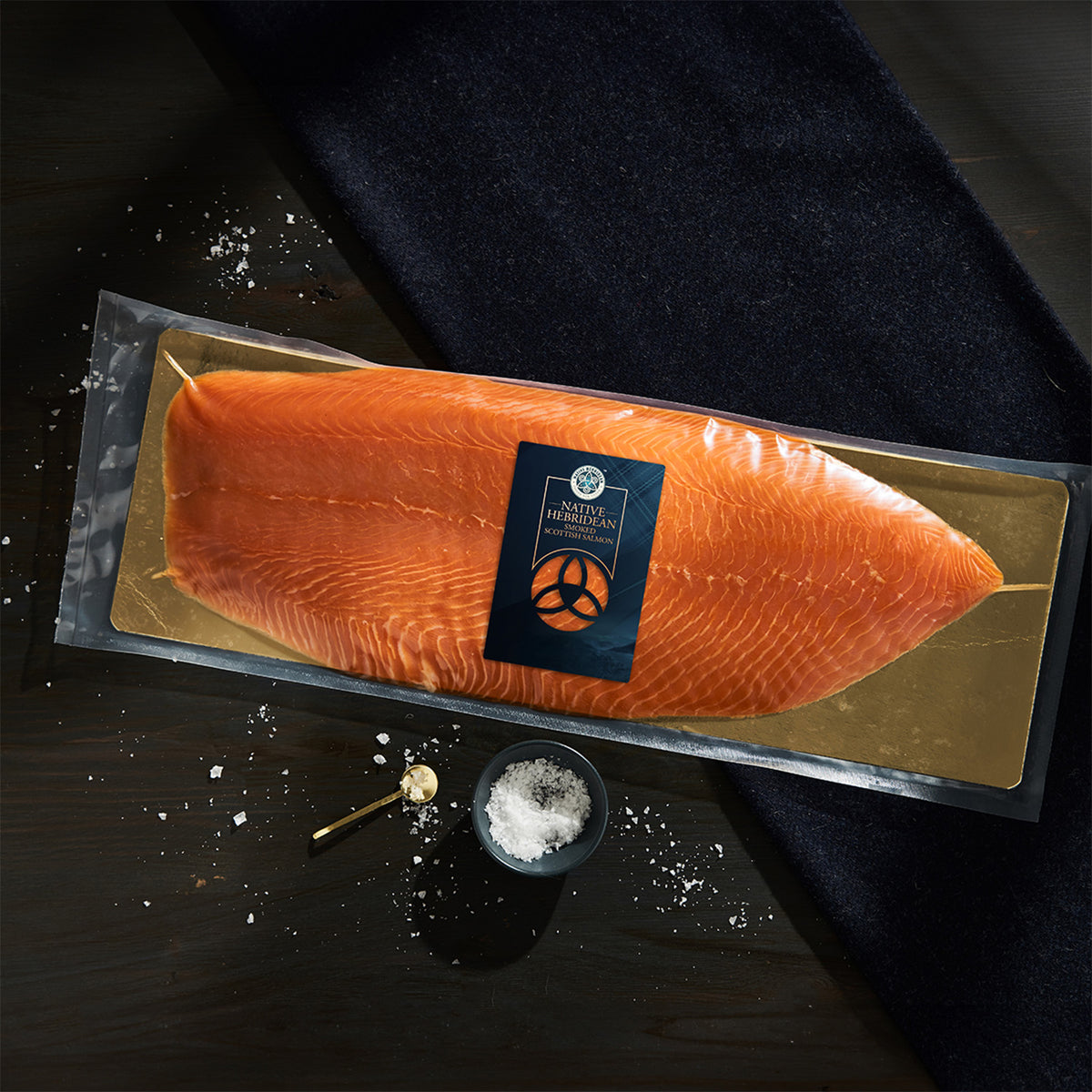 A vacuum-sealed package of salmon fillet with a gold foil tray and a central label, photographed on a dark wooden surface with scattered salt and a small bowl of salt nearby.